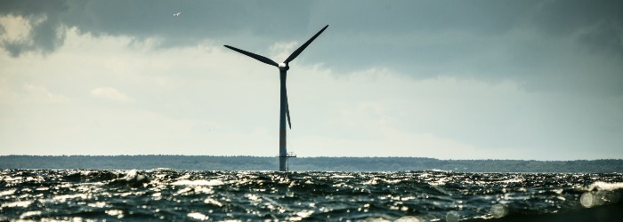 Offshore wind turbine standing in the ocean under a cloudy sky, with waves in the foreground reflecting sunlight.
