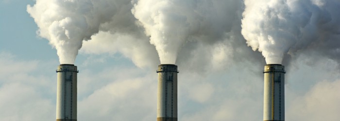 Three industrial smokestacks release thick white smoke into a cloudy sky, symbolizing air pollution and environmental impact.