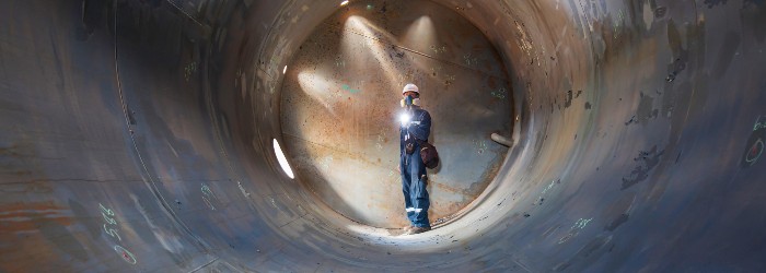 A worker in a hard hat stands inside a large metal cylinder, illuminating the space with a flashlight amid soft beams of light.