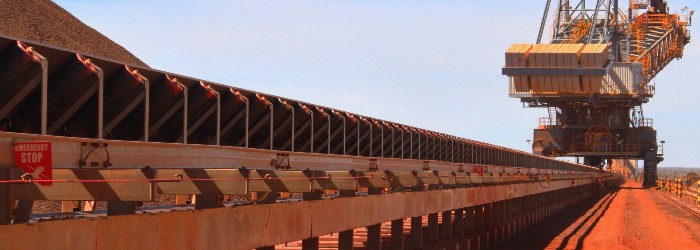 A conveyor belt system transports material, with a large overhead crane in a mining site under clear blue skies.