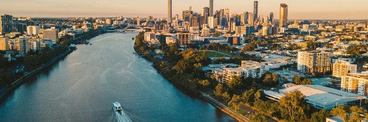 Aerial view of a river flowing through a vibrant cityscape at sunset, with a boat cruising along the water and skyscrapers in the background.