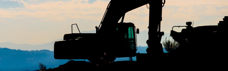 Silhouette of an excavator against a scenic backdrop of mountains and cloudy sky during sunset.