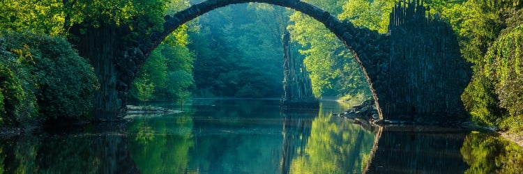A picturesque view of a stone arch bridge reflected in calm water, surrounded by lush greenery and trees.